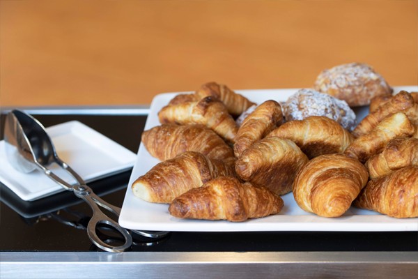 Croissants dusted with powdered sugar are served on a square, white plate. Silver serving tongs sit on a small plate alongside it.