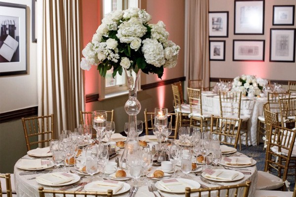 A tall arrangement of fresh-cut, white flowers sits at the center of a banquet table that is set for a formal sit down dinner.