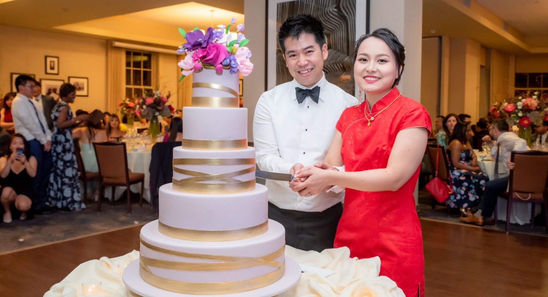 A happy couple about to cut into their cake, adorned with flowers and encased in gold ribbon.