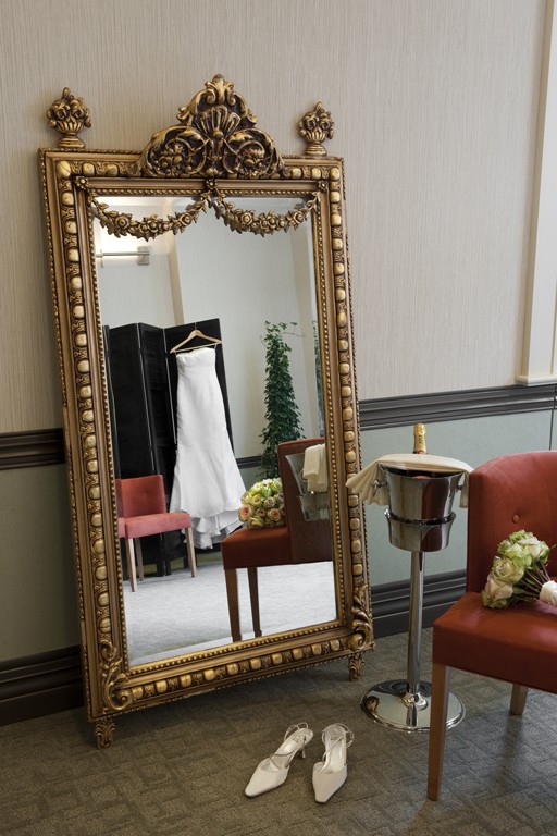 A wedding dress hangs next to a gilded mirror in the Faculty House Bridal Suite.