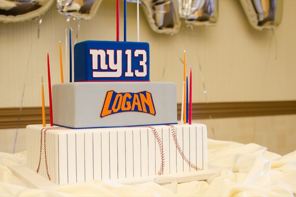 A three-tier cake is decorated with different New York sports themes. The bottom tier is covered with fondant and decorated to look like a baseball. The middle tier is gray and features the name 'Logan' in font reminiscent of the New York Knocks logo. The third tier is blue and reads, 'NY 13' in the style of the New York Giant logo.