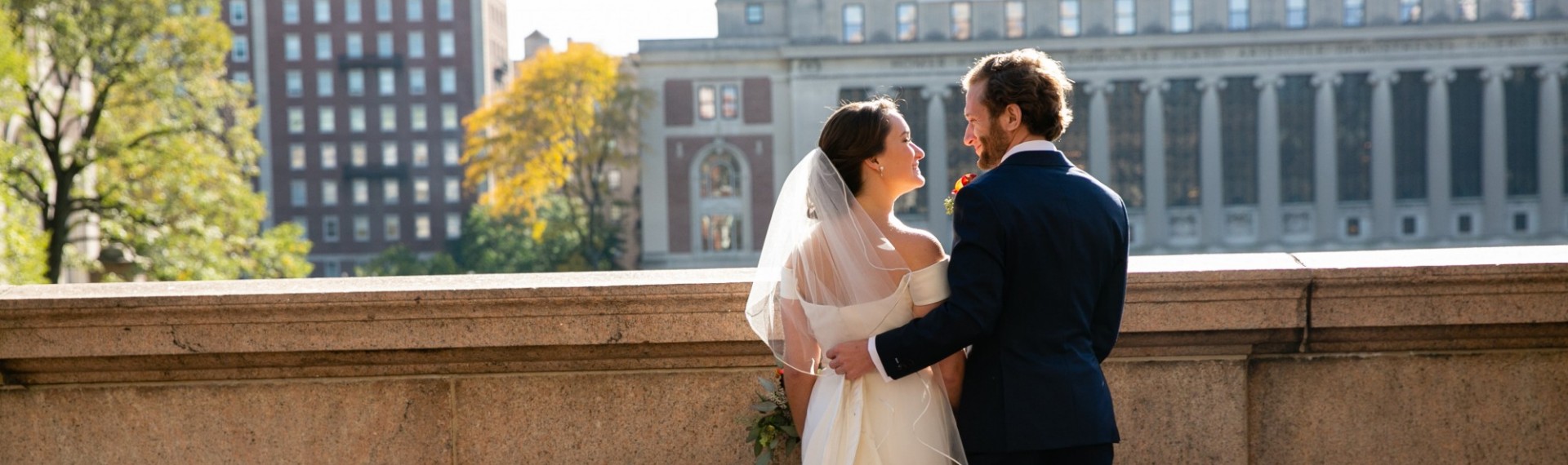 A bride and groom pause to smile at one another. Butler Library is visible in the background.