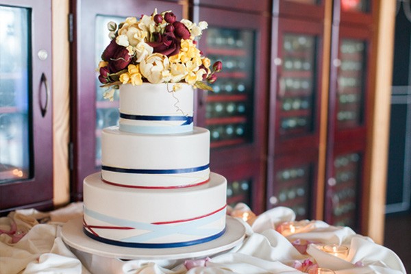 A three-tier cake is covered in white fondant. Each tier features bands of red and blue ribbon at its base. The cake is topped with a bouquet of ivory, yellow and burgundy sugar paste flowers.