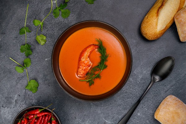 A bowl of lobster bisque is topped with a whole piece of claw meat with its shell removed. The bowl of bisque sits on a slate gray table and is surrounded by loaves of bread and fresh herbs.