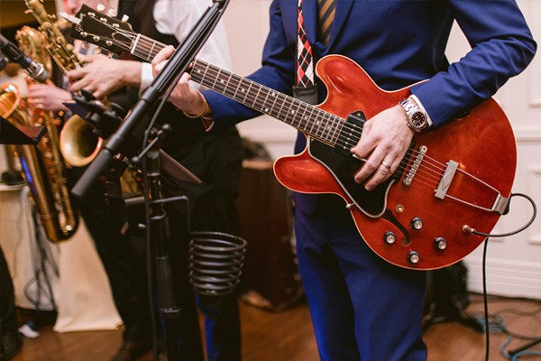 A musician wearing a blue pant suit plays the electric guitar. Musicians playing trumpets are visible in the background.