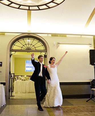 A newly-married couple enter their reception with their arms in the air, shouting.