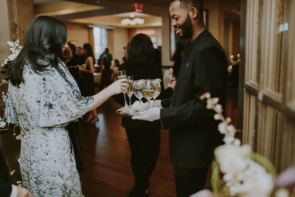 A person wearing a silver velvet gown takes a glass of sparkling wine from a tray offered by a member of the Faculty House wait staff