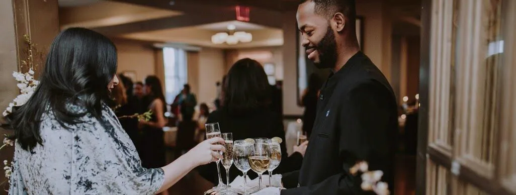 A guest wearing a silver velvet gown accepts a glass of champagne from tray offered by a Faculty House staff member wearing white gloves